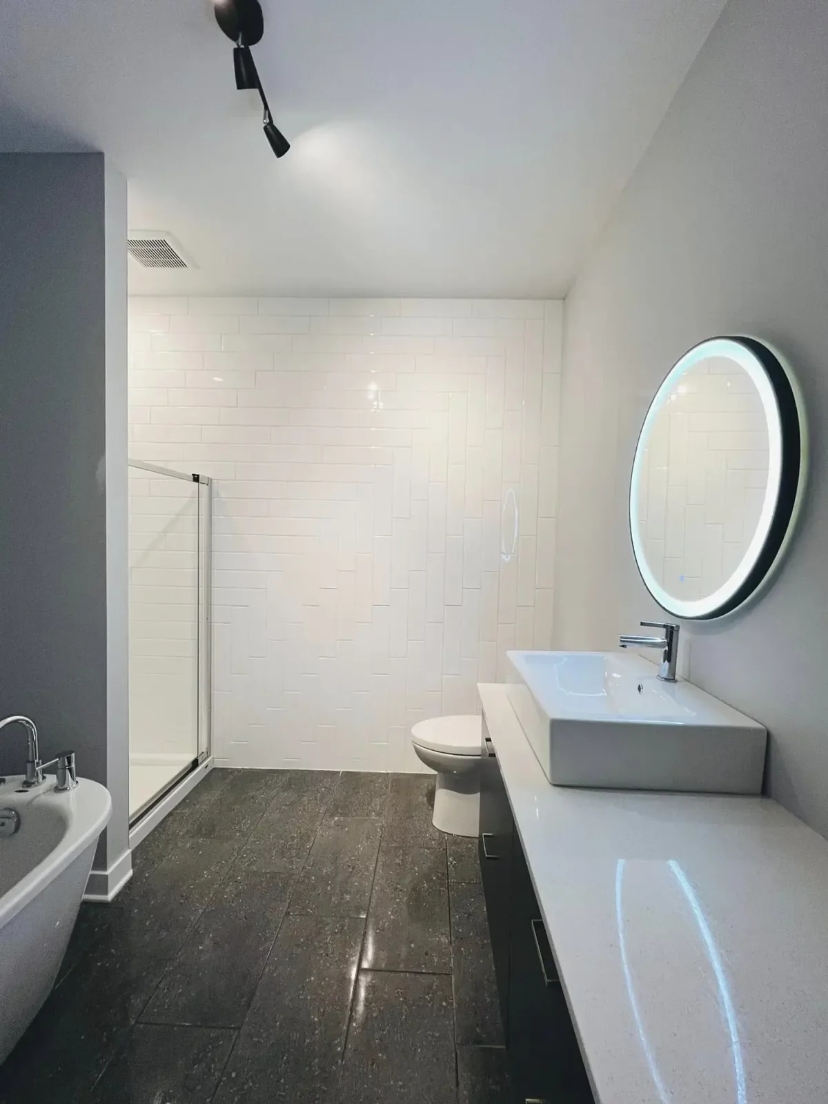 Salle de bain moderne avec douche vitrée, miroir DEL rond et plancher foncé – triplex luxueux Les Colibris Joliette